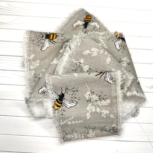 Op de afbeelding: Een grijs stoffen lint met een herhalend patroon van gele en zwarte hommels en witte bloemsilhouetten. Het lint heeft een geweven textuur en gerafelde, onafgewerkte randen langs de gehele lengte van het materiaal.