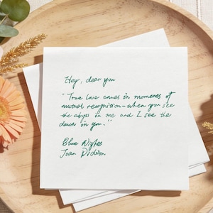 Puede incluir: Una pila de tarjetas cuadradas blancas con texto manuscrito verde, sobre una bandeja de madera. El texto incluye una cita de "Blue Nights" de Joan Didion. La bandeja está rodeada de flores y vegetación.