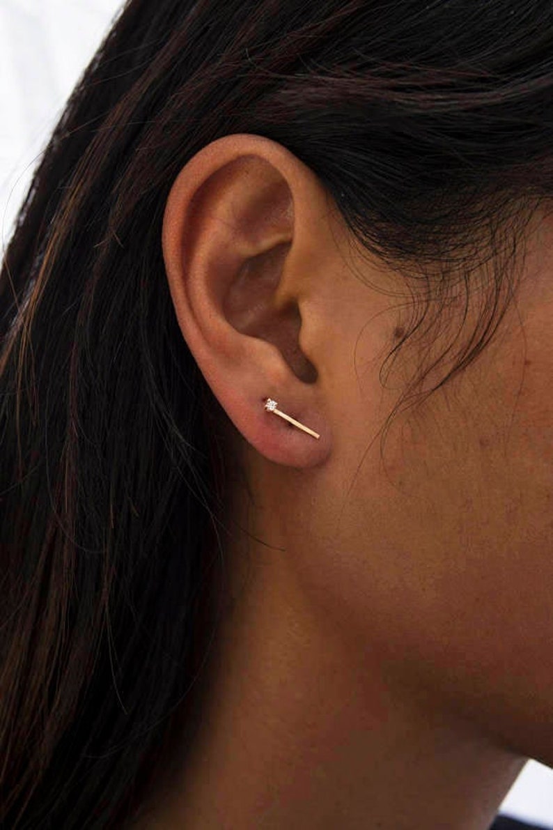 May include: A close-up of a person's ear with a small gold bar earring with a single diamond accent.