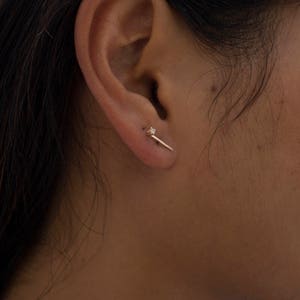 May include: A close-up of a person's ear with a small gold bar earring with a single diamond accent.