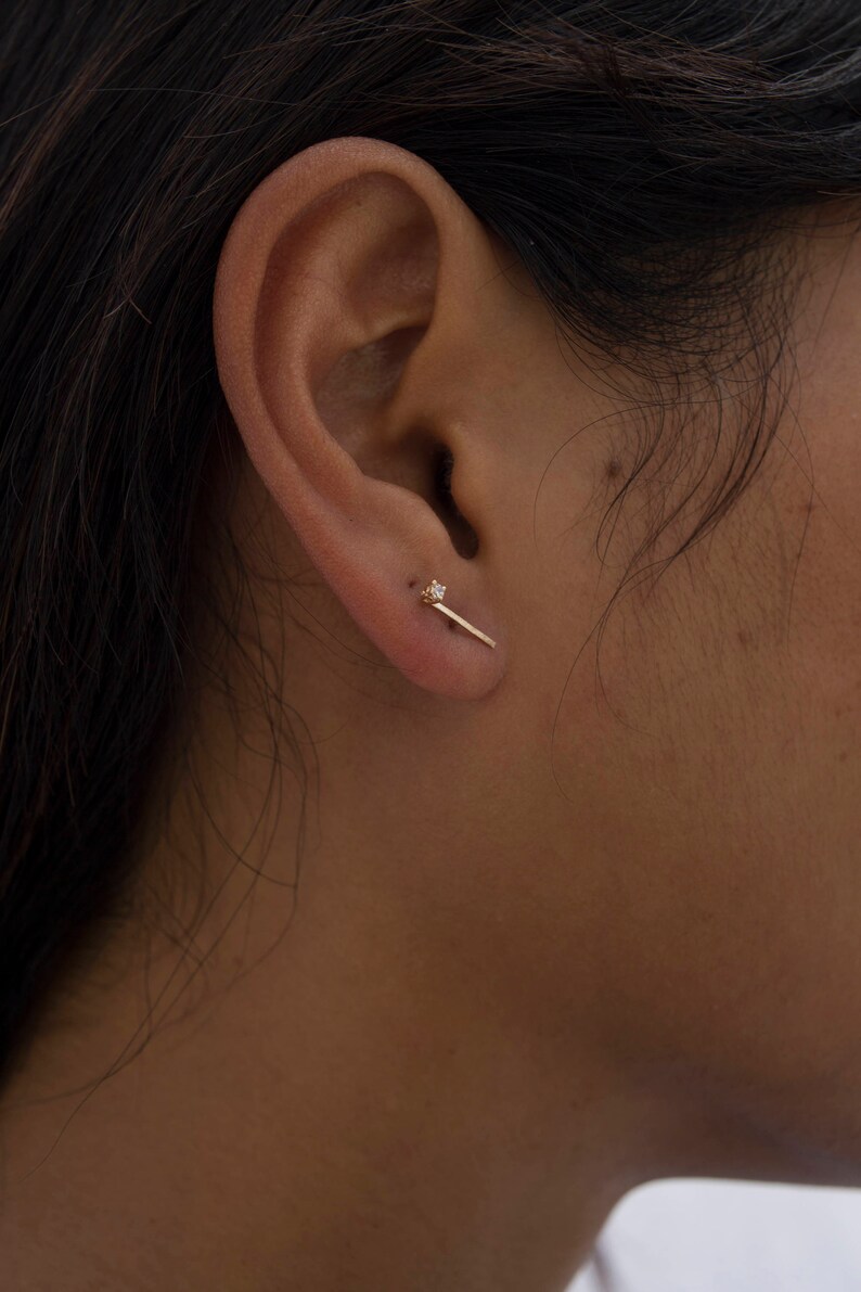 May include: A close-up of a person's ear with a small gold bar earring with a single diamond accent.