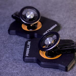 Peut inclure: Deux supports de téléphone de voiture noirs et argentés. Chacun a un design circulaire avec un centre argenté et un anneau extérieur noir. La base est noire avec un cercle orange. Le mot PARALLAX est imprimé.