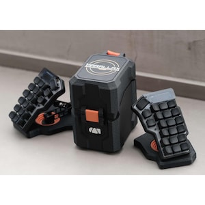 May include: Black and orange mechanical keyboard with a unique design. The keyboard has a split layout with individual keycaps and a central storage unit. The word "PARALLAX" is printed on the top of the storage unit.