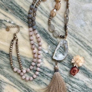 May include: A long necklace featuring a mix of faceted champagne-colored beads, smooth grey beads, and pale pink round beads. The piece includes a large, irregular white geode pendant with a gold-toned edge and a long, beige fabric tassel.
