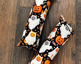 May include: Two Halloween-themed cat toys. The toys are rectangular and feature a black background with white ghosts, orange pumpkins, and candy corn. The cats are dressed in ghost costumes. The toys are on a dark wood surface.