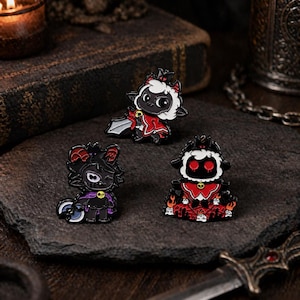 Op de afbeelding: Drie emaille pins met cartoon lammeren in verschillende poses. De pins zijn voornamelijk zwart, rood en wit, met details zoals kronen, capes en zwaarden. De pins zijn geplaatst op een donker, getextureerd oppervlak.