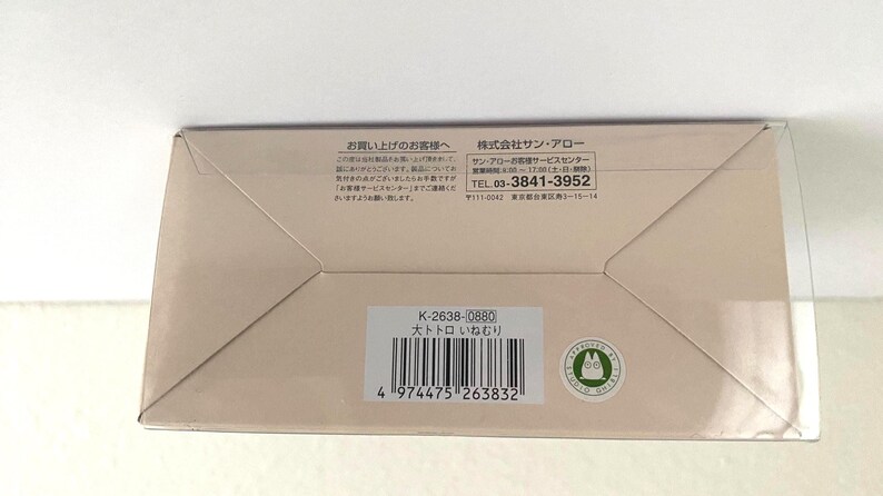 May include: A beige rectangular box with Japanese text and a barcode. The box has a clear plastic top and a green circular sticker with a cartoon character. The text includes a phone number and the words "大トトロ いねむり".