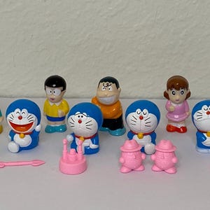 May include: A collection of colorful Doraemon figurines and accessories. The figures include Doraemon, Nobita, and other characters. There are also pink accessories, including a crown, hats, and a small arrow. The figurines are on a white surface.