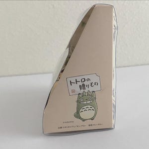 May include: A light beige box featuring a cartoon Totoro character holding a sign with Japanese text. The box has a triangular shape and is decorated with additional Japanese text and illustrations.