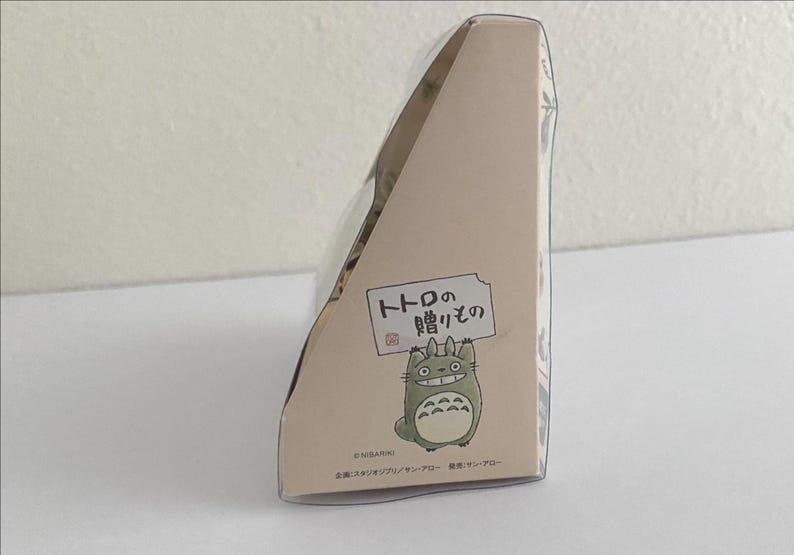 May include: A light beige box featuring a cartoon Totoro character holding a sign with Japanese text. The box has a triangular shape and is decorated with additional Japanese text and illustrations.