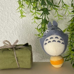 May include: A plush, gray Totoro character with a white belly and a yellow and orange ball. Beside it is a small, olive green fabric package tied with twine. Green foliage is in the background.