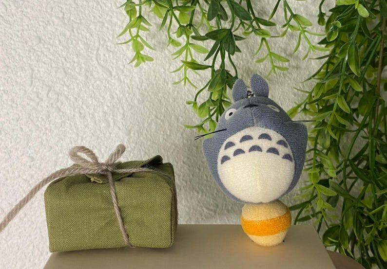 May include: A plush, gray Totoro character with a white belly and a yellow and orange ball. Beside it is a small, olive green fabric package tied with twine. Green foliage is in the background.