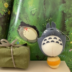 May include: A plush Totoro character with a white belly and a yellow and orange ball. A small green gift box tied with twine sits to the left. The background features a colorful illustration of a forest scene with Totoro.