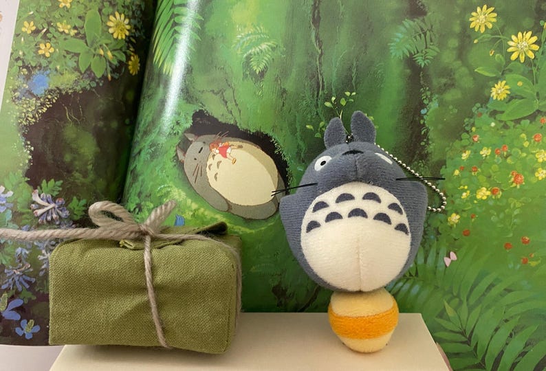 May include: A plush Totoro character with a white belly and a yellow and orange ball. A small green gift box tied with twine sits to the left. The background features a colorful illustration of a forest scene with Totoro.