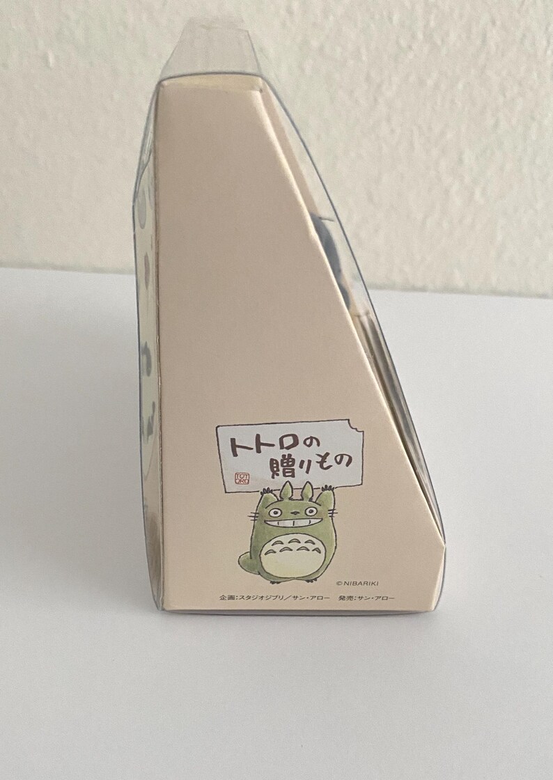 May include: A beige cardboard box with a clear plastic top. The box features a cartoon illustration of Totoro and Japanese text. The box is a gift item.