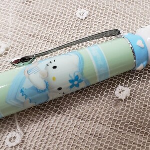 May include: A light green and blue ballpoint pen featuring a Hello Kitty character design. The pen barrel displays a printed graphic of Hello Kitty in a blue outfit, while the top cap is topped with a three-dimensional Hello Kitty figure.