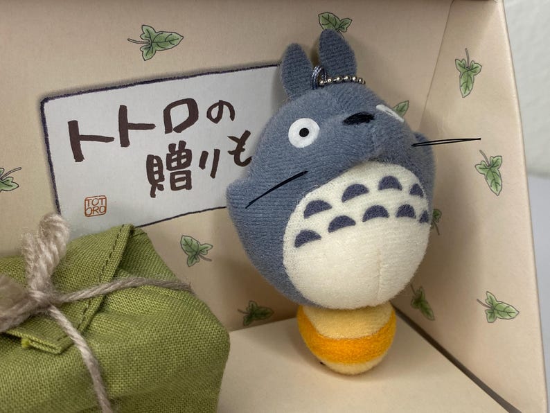 May include: A plush Totoro keychain in a gift box. The gray Totoro has a white belly and a yellow base. The box has Japanese text and leaf illustrations. A green fabric package is tied with twine.