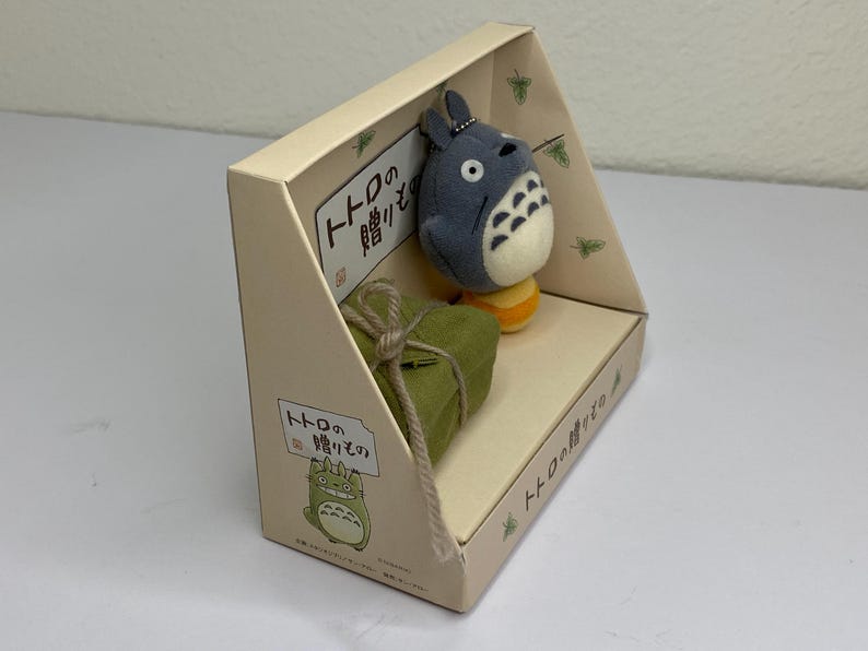 May include: A beige cardboard display box featuring a plush Totoro character and a small green gift box tied with twine. The box has Japanese text and a cartoon illustration of Totoro.