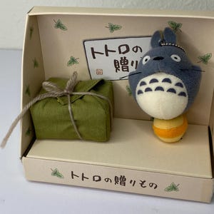May include: A boxed set featuring a plush Totoro character with a yellow and orange base, and a wrapped green package tied with twine. The box has Japanese text and leaf illustrations.