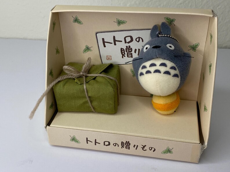 May include: A boxed set featuring a plush Totoro character with a yellow and orange base, and a wrapped green package tied with twine. The box has Japanese text and leaf illustrations.