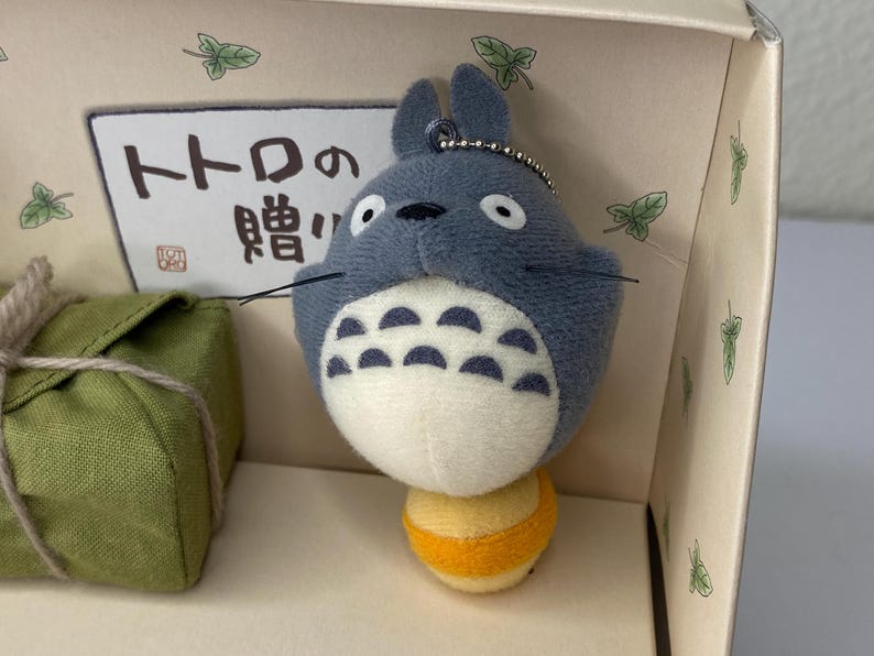 May include: A plush Totoro keychain in a box. The gray Totoro has a white belly with black markings and a yellow and orange base. The box has Japanese text and green leaf accents. A green package with a rope tie is also visible.