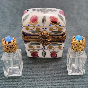 May include: A white porcelain trinket box with a hinged lid, decorated with pink roses and gold accents. Two small, clear glass perfume bottles with ornate gold filigree caps and blue cabochon tops are on either side.