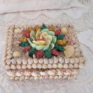 May include: A rectangular box decorated with small, white seashells. The lid features a large, colourful seashell flower in shades of green, yellow, and red. The box sits on a white, lace-covered surface.