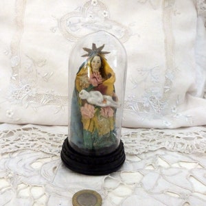 Puede incluir: Figura religiosa dentro de una cúpula de cristal. La figura representa a una mujer con una túnica amarilla y azul, sosteniendo un objeto blanco y rodeada de flores rosas. La cúpula descansa sobre una base negra.