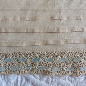 Può includere: Un tessuto beige con un bordo in pizzo. Il pizzo è di colore crema con accenti di nastro blu.