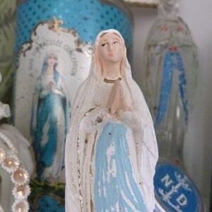 May include: A white figurine of the Virgin Mary with a blue robe and a white veil. The figurine is standing on a table with other religious items.
