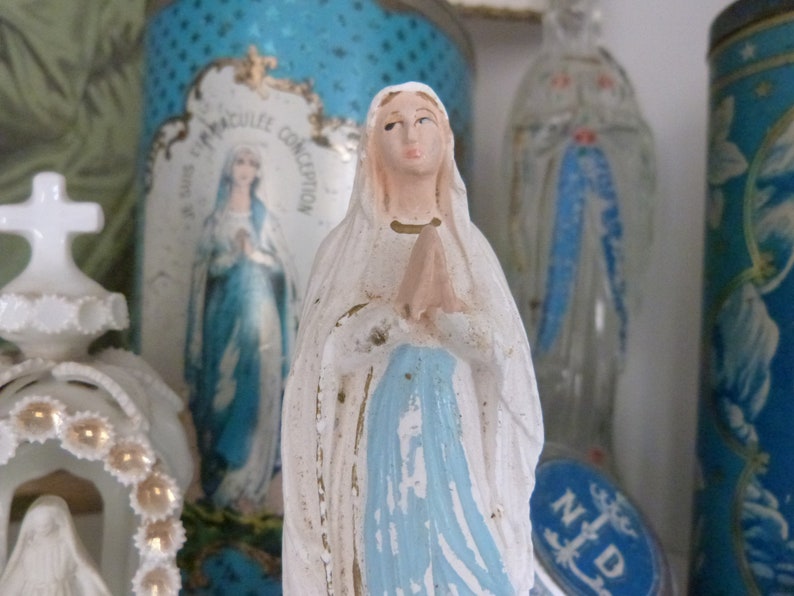 May include: A white figurine of the Virgin Mary with a blue robe and a white veil. The figurine is standing on a table with other religious items.