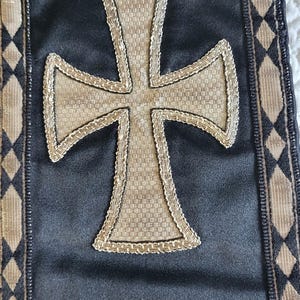 May include: Black textile with a large, textured cross in the center. The cross is beige and outlined with a gold-colored trim. The edges of the textile have a decorative border with diamond patterns.