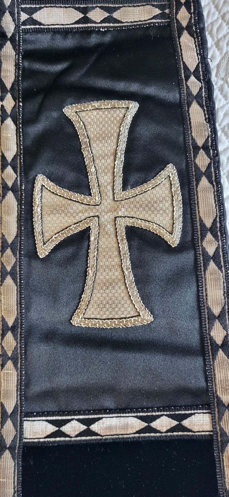 May include: Black textile with a large, textured cross in the center. The cross is beige and outlined with a gold-colored trim. The edges of the textile have a decorative border with diamond patterns.