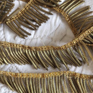 May include: Close-up of gold-colored fringe trim. The trim features rows of tightly coiled, spring-like elements hanging from a woven base. The fringe is arranged in a repeating pattern, creating a textured and decorative element. The background is a white, textured fabric.