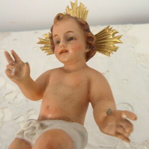 May include: A vintage figurine of a baby with a golden halo. The baby has light skin, brown hair, and is wearing a white diaper. The figurine is posed with one hand making a peace sign.