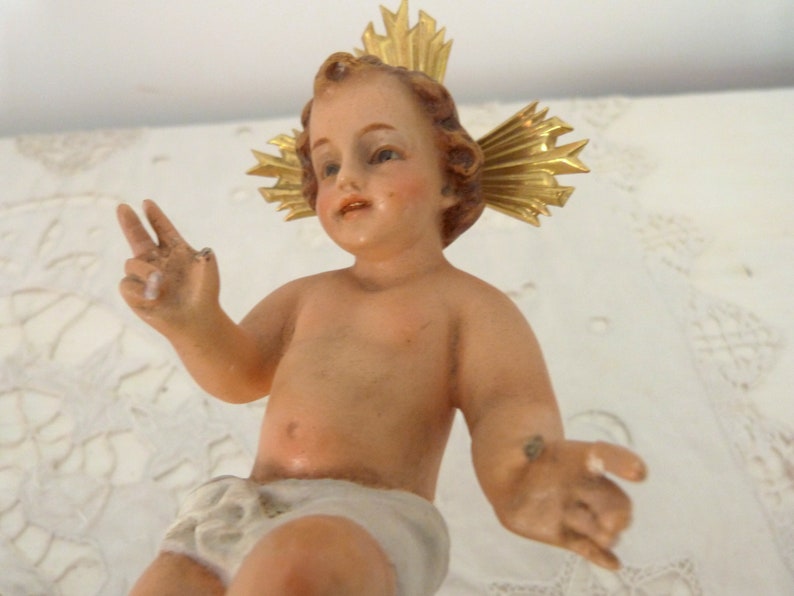 May include: A vintage figurine of a baby with a golden halo. The baby has light skin, brown hair, and is wearing a white diaper. The figurine is posed with one hand making a peace sign.