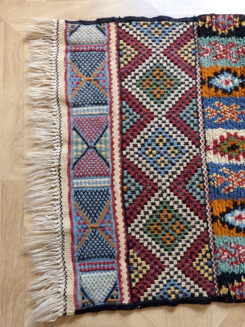 Op de afbeelding: Een kleurrijk geweven tapijt met een geometrisch patroon in tinten rood, blauw, groen en geel. Het tapijt heeft aan &eacute;&eacute;n kant franjes.