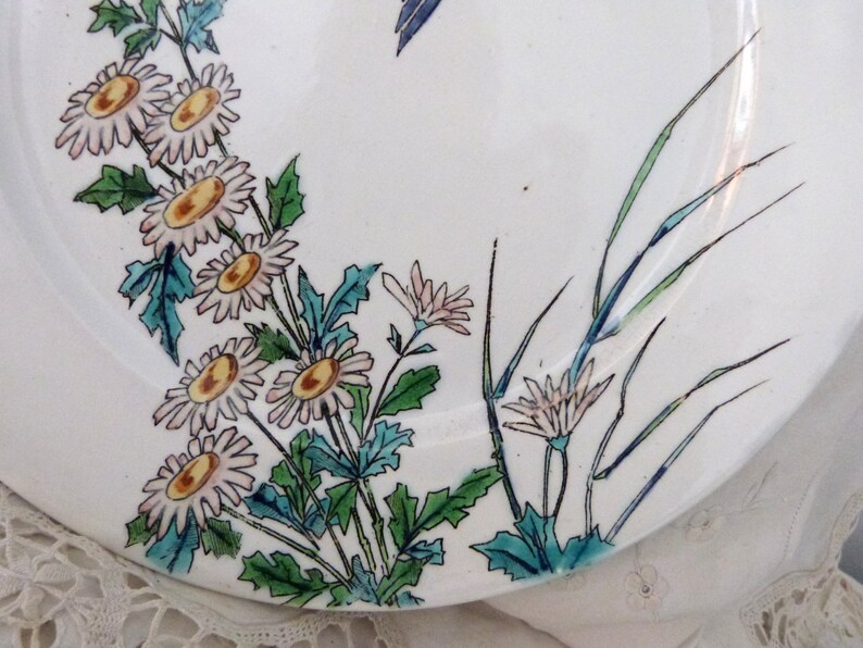 Pode incluir: Um prato de cer&acirc;mica branco com um design floral. O prato apresenta flores rosa e brancas com centros amarelos, folhas verdes e caules azuis. O design lembra um estilo japon&ecirc;s.