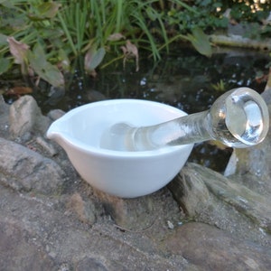 May include: A white ceramic mortar and pestle with a clear glass pestle. The mortar and pestle are sitting on a rocky surface.
