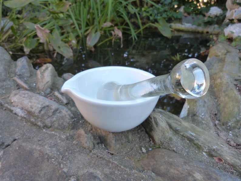 May include: A white ceramic mortar and pestle with a clear glass pestle. The mortar and pestle are sitting on a rocky surface.