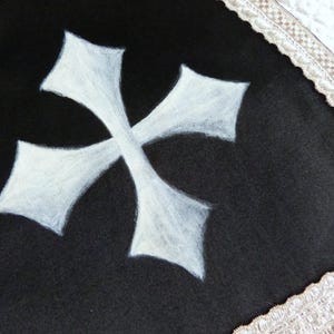Może przedstawiać: Czarny materiał z białym wzorem krzyża maltańskiego. Krzyż jest wyśrodkowany i wygląda na ręcznie malowany. Krawędź materiału jest obszyta kremową, dekoracyjną bordiurą.