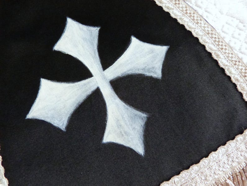 Może przedstawiać: Czarny materiał z białym wzorem krzyża maltańskiego. Krzyż jest wyśrodkowany i wygląda na ręcznie malowany. Krawędź materiału jest obszyta kremową, dekoracyjną bordiurą.