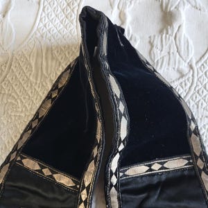 May include: Black velvet scarf with silver geometric trim. The scarf is folded, revealing its interior. The trim features a repeating pattern of diamonds and lines. The scarf is resting on a white, patterned surface.