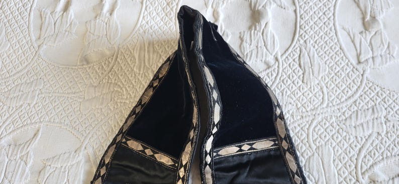 May include: Black velvet scarf with silver geometric trim. The scarf is folded, revealing its interior. The trim features a repeating pattern of diamonds and lines. The scarf is resting on a white, patterned surface.
