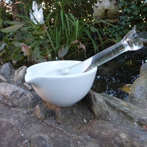 May include: A white ceramic mortar and pestle set with a clear glass pestle. The mortar and pestle are sitting on a rock surface.