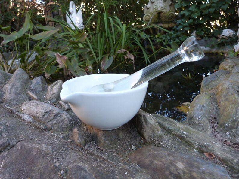 May include: A white ceramic mortar and pestle set with a clear glass pestle. The mortar and pestle are sitting on a rock surface.