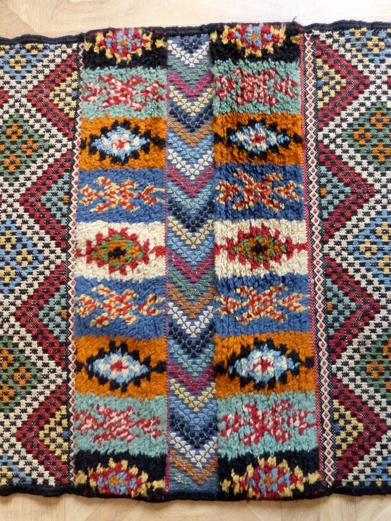 Op de afbeelding: Een kleurrijk Marokkaans tapijt met geometrische patronen in tinten rood, blauw, oranje en groen. Het tapijt heeft een dikke, textuurrijke pool en is geweven met ingewikkelde details.