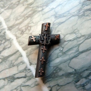 May include: A black metal cross with a silver fleur-de-lis design and a copper-colored center. The cross is decorated with smaller fleur-de-lis accents.