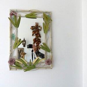 May include: A white metal mirror with a decorative frame featuring green leaves and pink flowers. The mirror reflects a person holding a camera.