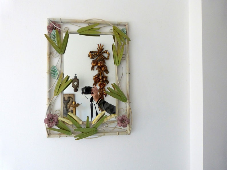 May include: A white metal mirror with a decorative frame featuring green leaves and pink flowers. The mirror reflects a person holding a camera.